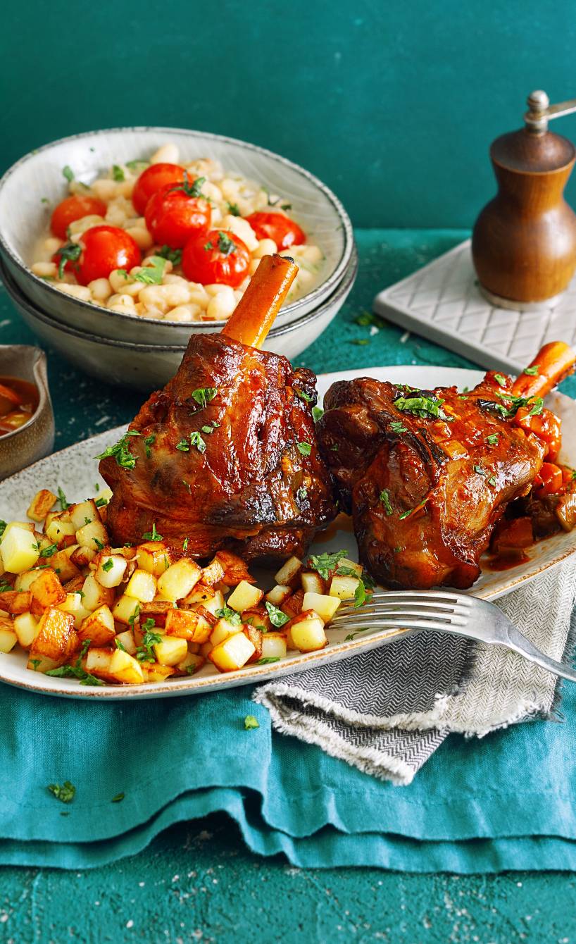 Zwei gebackene Lammhaxen auf einem Teller angerichtet mit knsuprigen Kartoffelwürfeln, daneben eine Schale mit Bohnen als Beilage