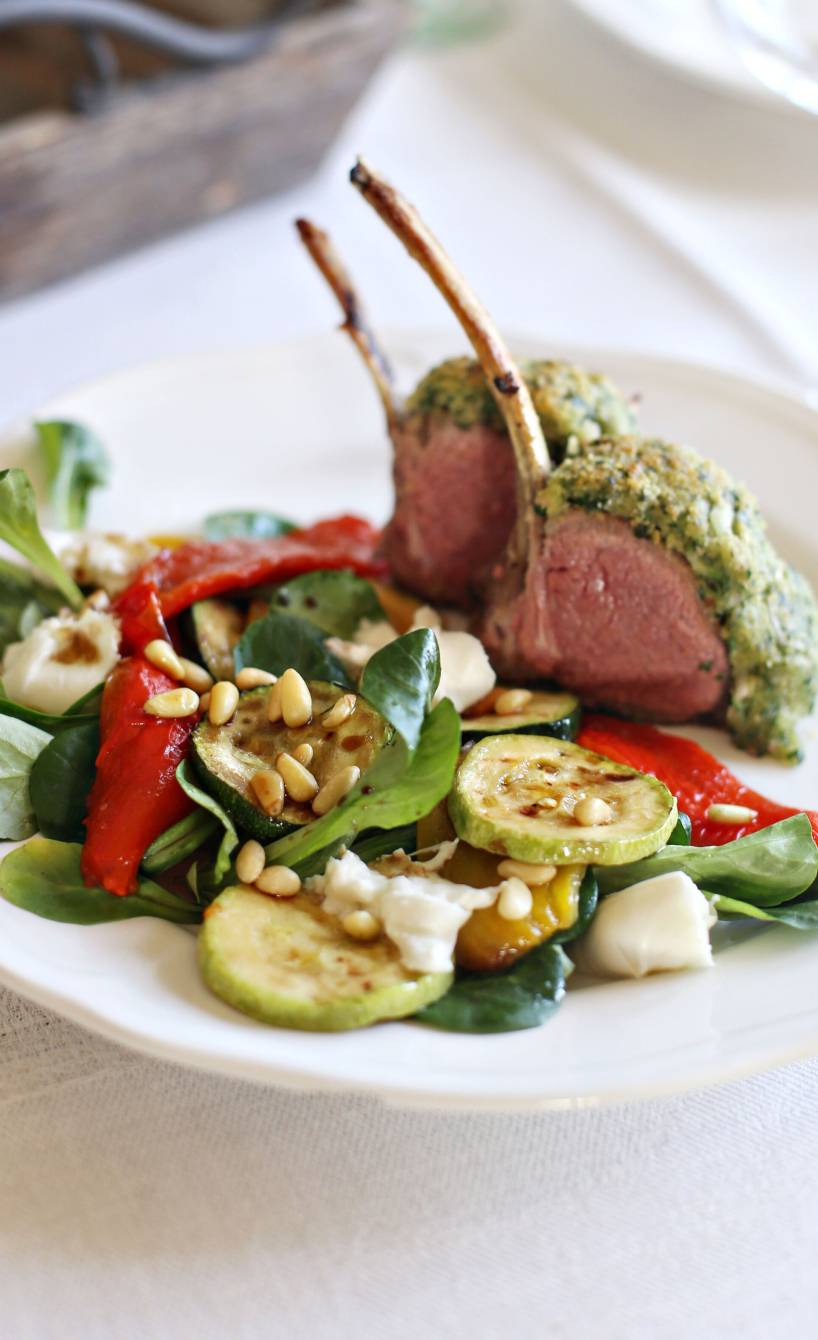 Lammkarree mit Parmesankruste angerichtet auf Antipastisalat getoppt von Pinienkernen auf einem Teller