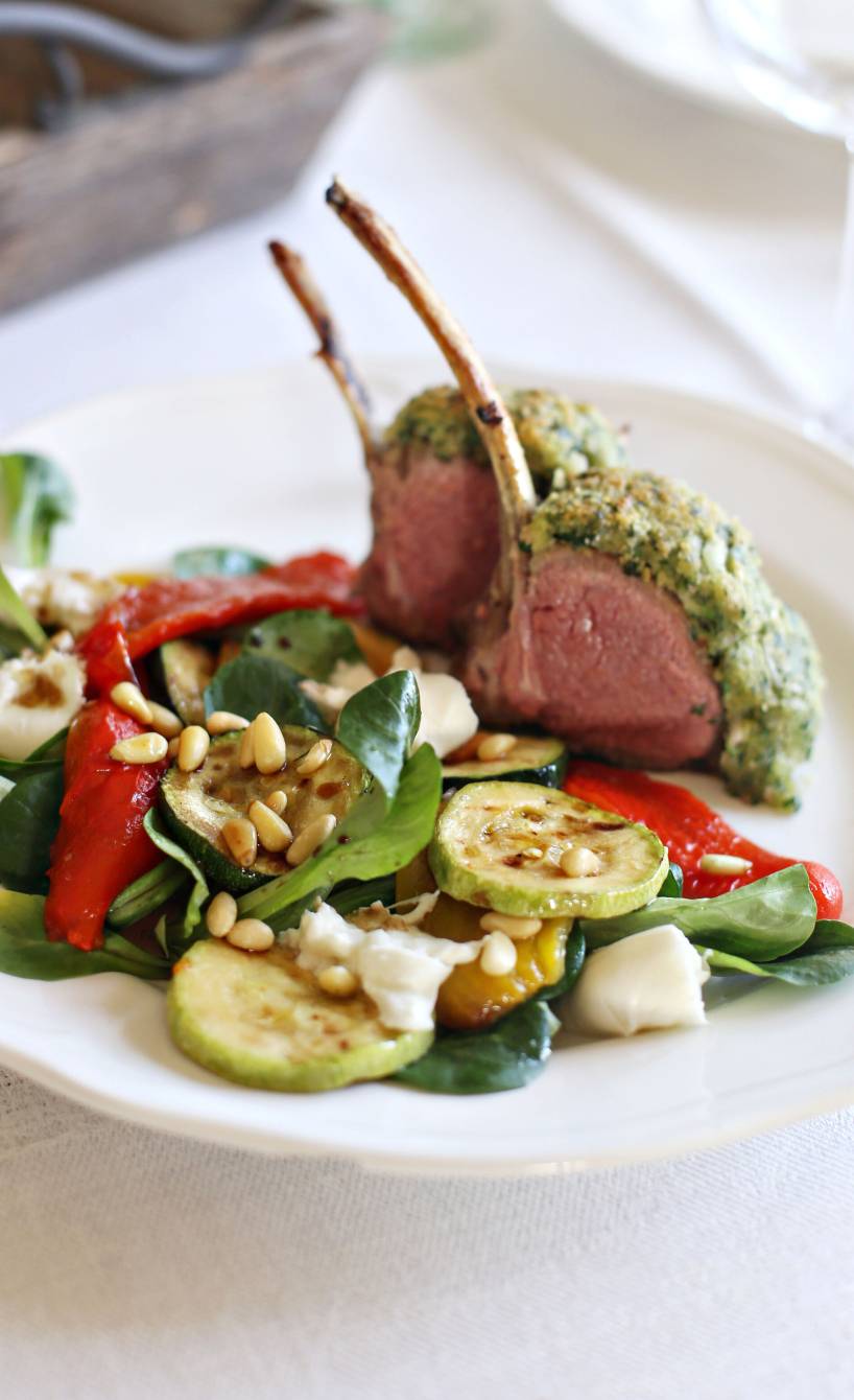 Lammkarree mit Parmesankruste angerichtet auf Antipastisalat getoppt von Pinienkernen auf einem Teller
