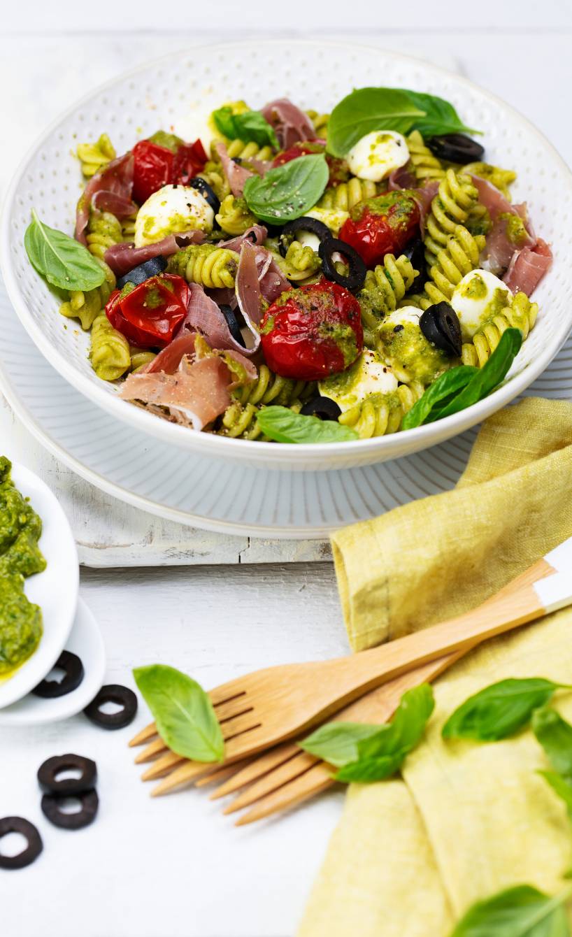 Mediterraner, lauwarmer Nudelsalat mit Olivenpesto, Schinken und Ofentomaten angerichtet auf einem tiefen Teller