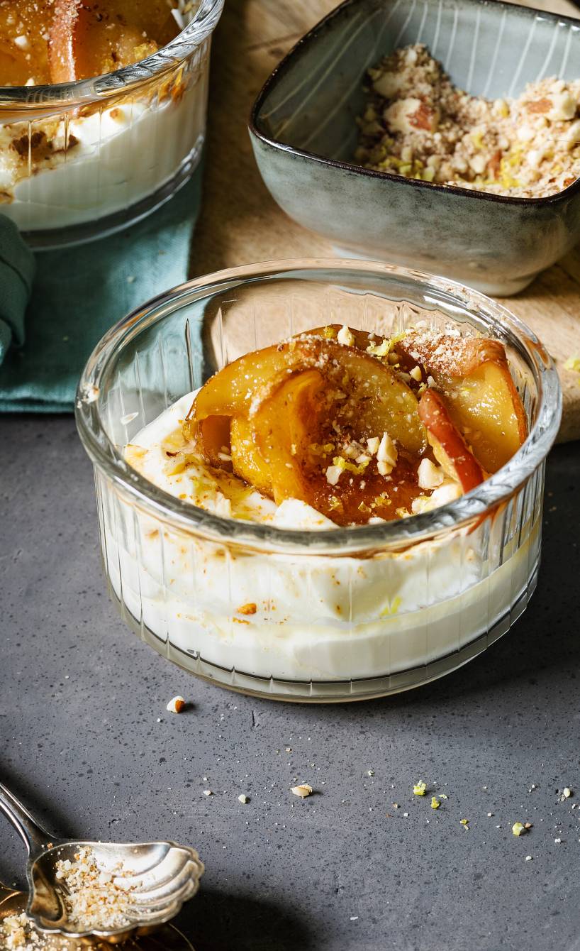 Limoncello-Mascarponedessert getoppt mit karamelliiserten Apfelspalten und Zitronen-Bröseln angerichtet in Dessertgläsern auf einem Tisch