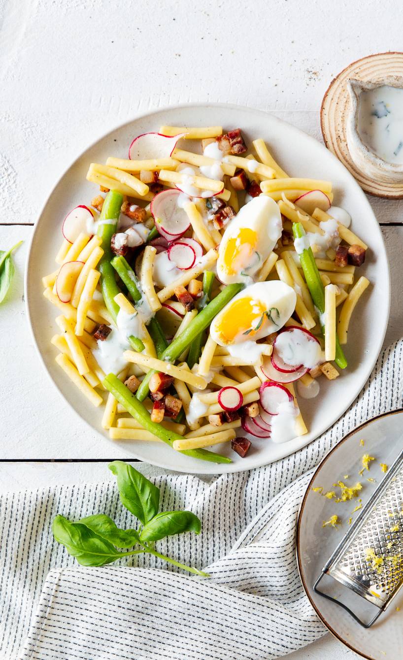 Nudelsalat aus Maccheroni mit Pancetta, Ei und grünen Bohnen auf einem Teller, daneben Joghurtdressing