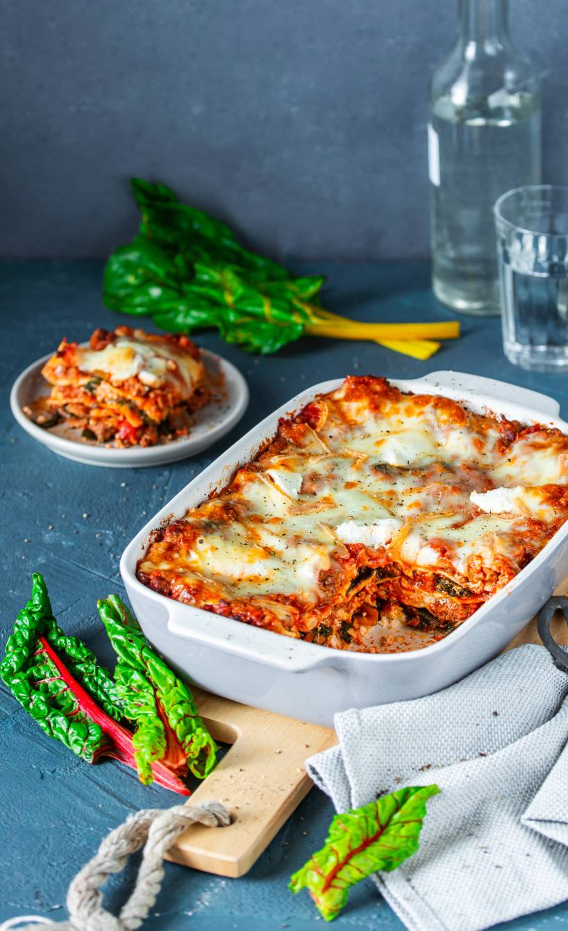 Mangoldlasagne mit Hackfleisch angeschnitten in einer Auflaufform, daneben Mangoldblätter