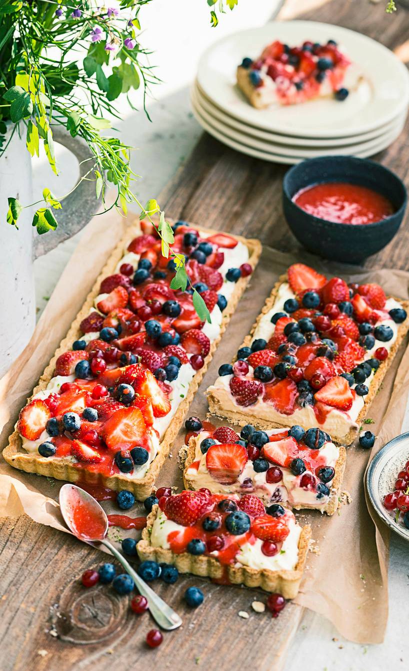 Zwei gebackene Beeren-Tartes mit Mascarpone-Joghurt-Creme getoppt von frischen Beeren angerichtet auf einem Holztisch