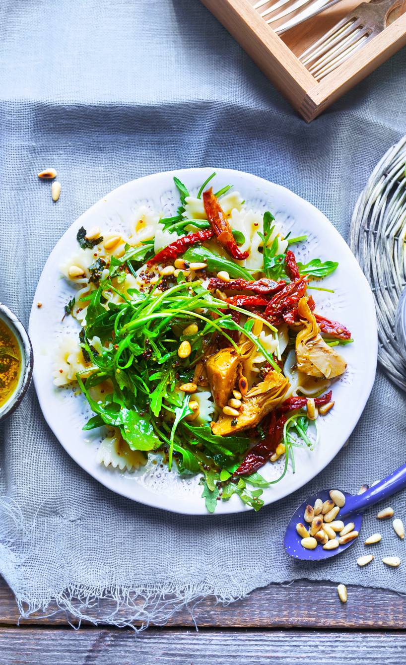 Mediterraner Nudelsalat auf einem Teller bestreut mit Pinienkernen, daneben eine Schale mit Vinaigrette