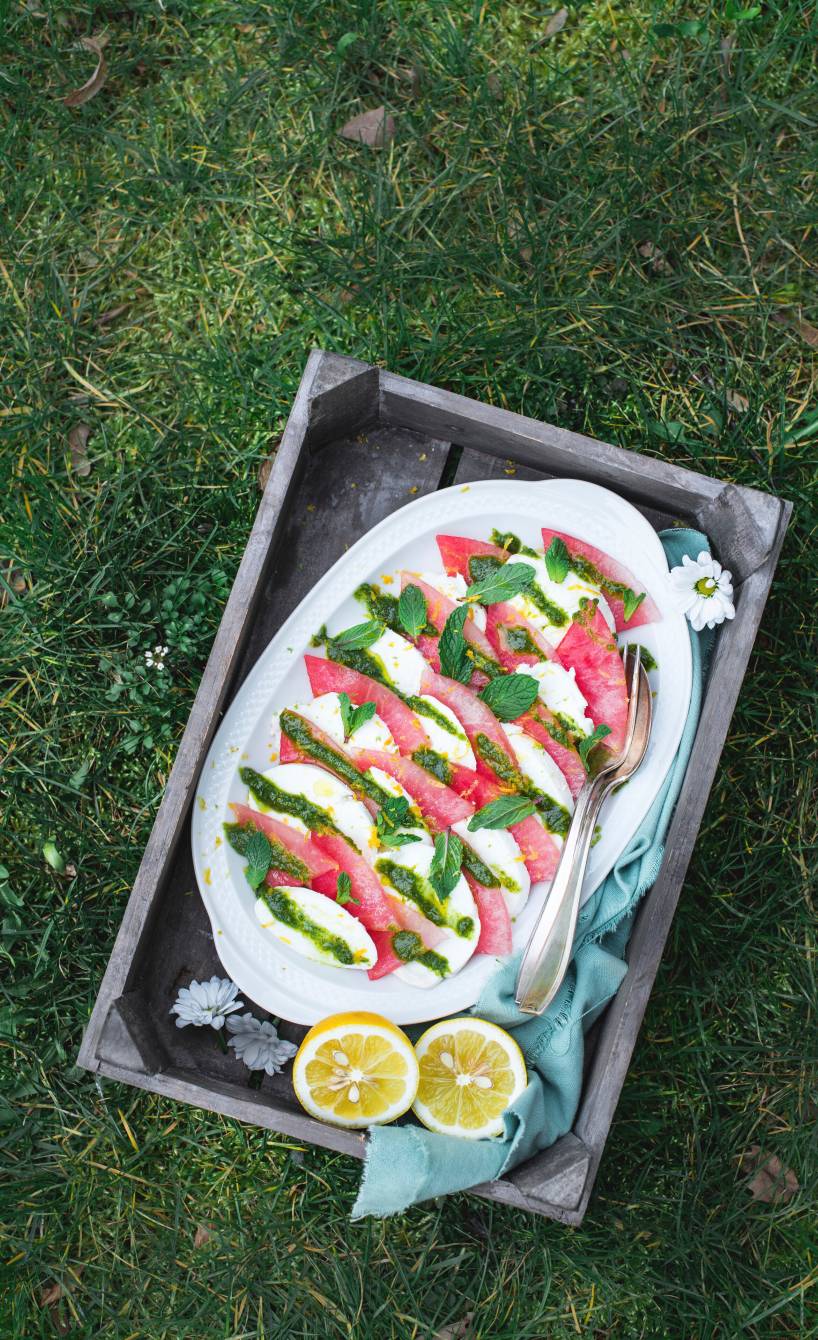 Caprese-Salat angerichtet auf einer weißen Platte aus Melonen- und Mozzarella-Streifen getoppt mit Pesto und Minze