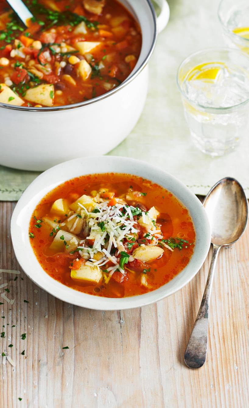 Minestrone in einer Suppenschüssel getoppt von Patmesanspänen, daneben ein voller Suppentop mit einer Kelle darin