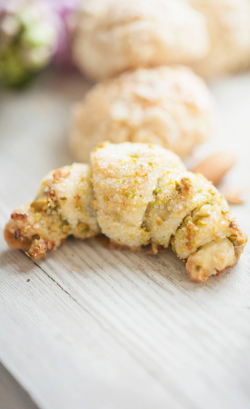 Ein Vanille-Hörnchen aus Frischkäseteig mit Pistazienfüllung und -topping auf einem hellen Holztisch