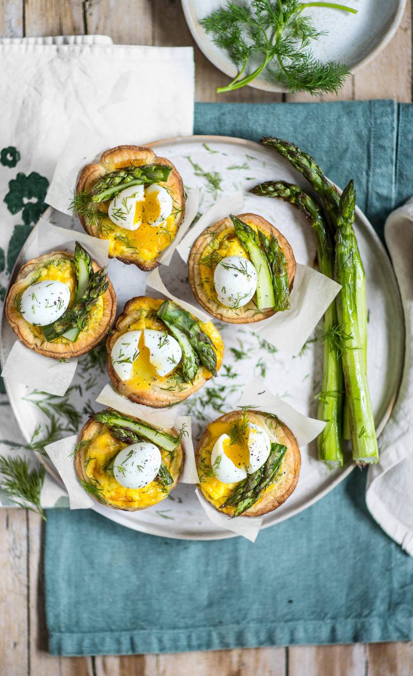 Mehrere selbstgebackene Mini-Spargel-Quiches aus Blätterteig mit Wachteleiern angerichtet auf einer Servierplatte