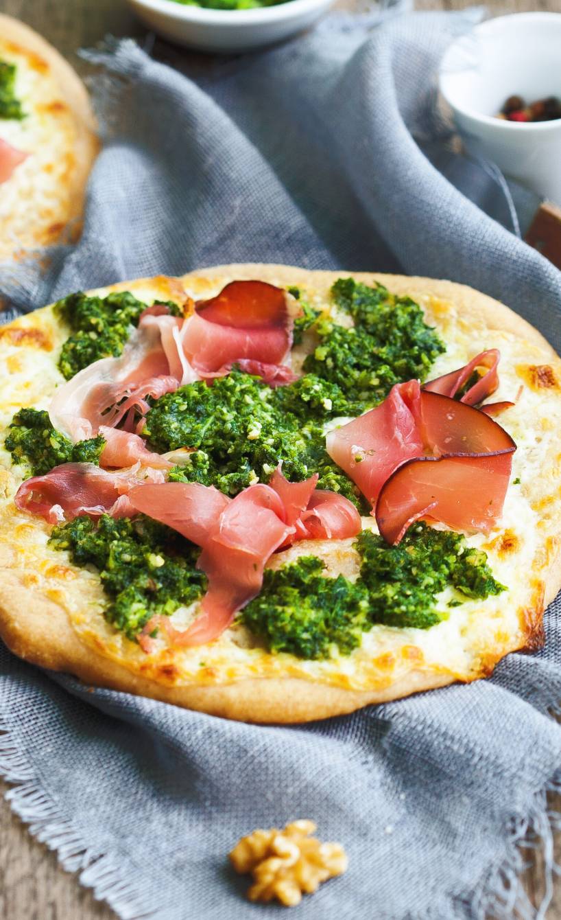 Zwei Minipizzen auf einem Holztisch mit Grünkohlpesto und Speck, daneben Walnusskerne
