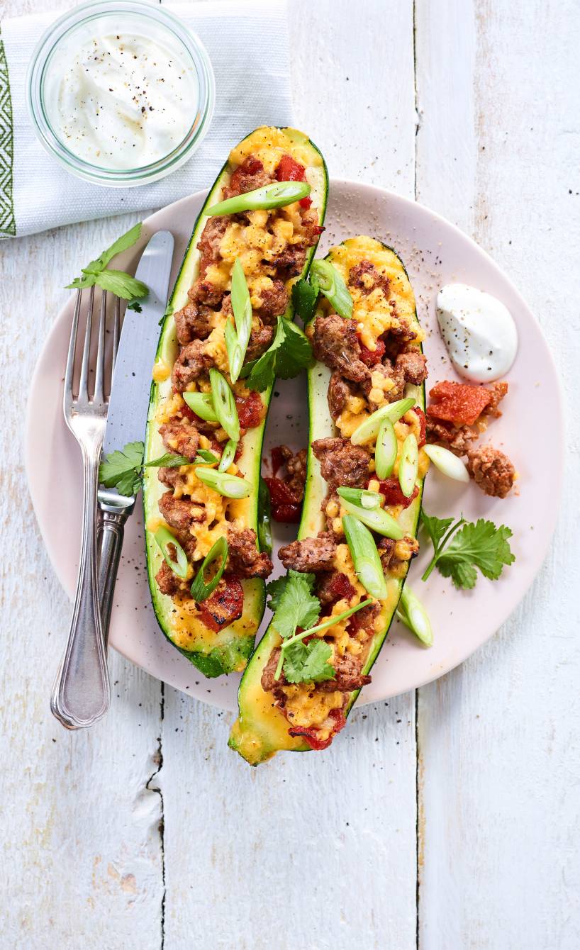 Zwei halbe, gebackene Zucchini gefüllt mit Hackfleisch à la Bolognese und mit Mozzarella überbacken angerichtet auf einem Teller mit Joghurtdip
