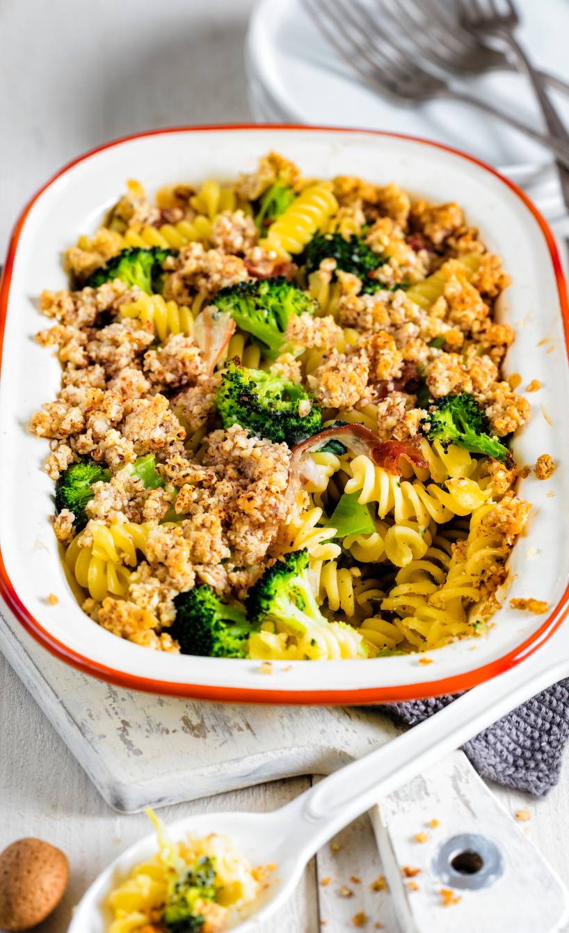 Nudelgratin mit Brokkoli, Pancetta und Mandelhaube in einer Auflaufform auf einem weißen Holztisch