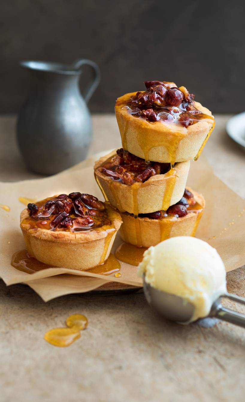 Vier karamellisierte Honig-Nuss-Törtchen mit Orangenguss angerichtet auf einem Dessertteller, daneben ein Eisportionierer mit einer Kugel Vanilleeis