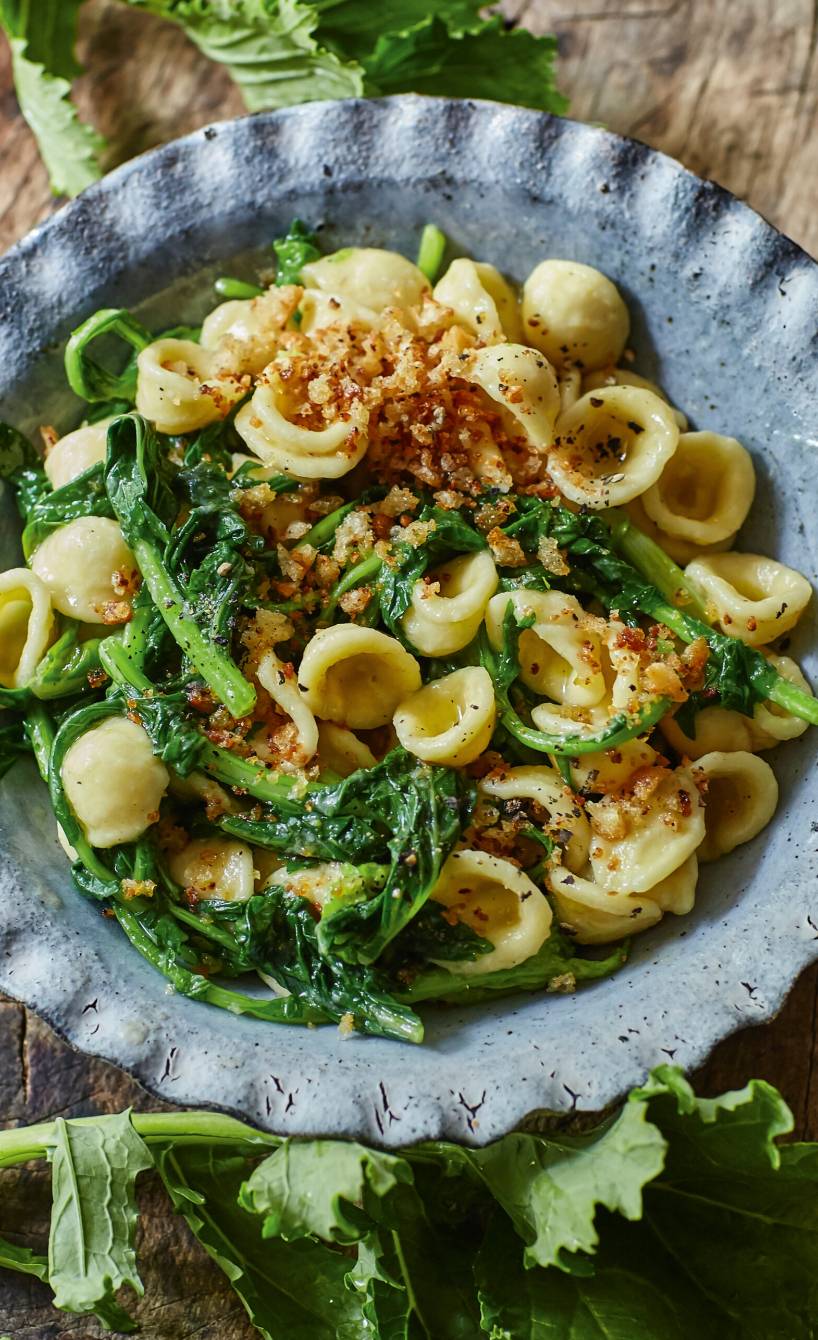 Orecchiette mit Cime di Rapa angerichtet auf einem Teller, daneben Stängelkohl auf dem Tisch