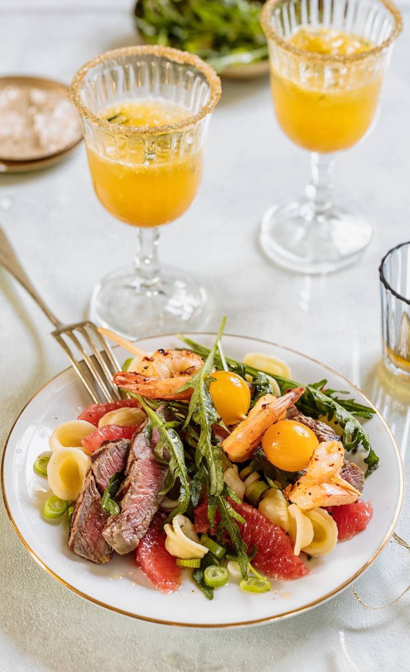 Nudelsalat mit Steakstreifen, Garnelenspießen und Grapefruitstücken auf einem Teller, daneben Dressing in einem Glas
