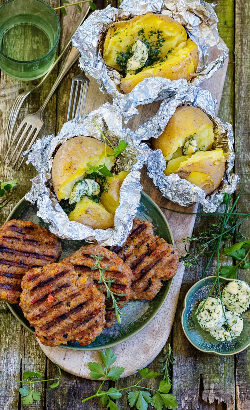 Mehrere Frikadellen mit Paprika und Mozzarella neben Backkartoffeln aus der Pfanne angerichtet auf einem Holzbrett
