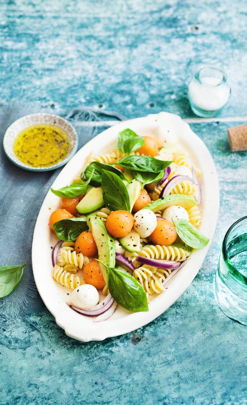 Pastasalat mit Melonenbällchen und Mozzarellakugeln auf einem ovalen Teller, darauf Avocadostücke