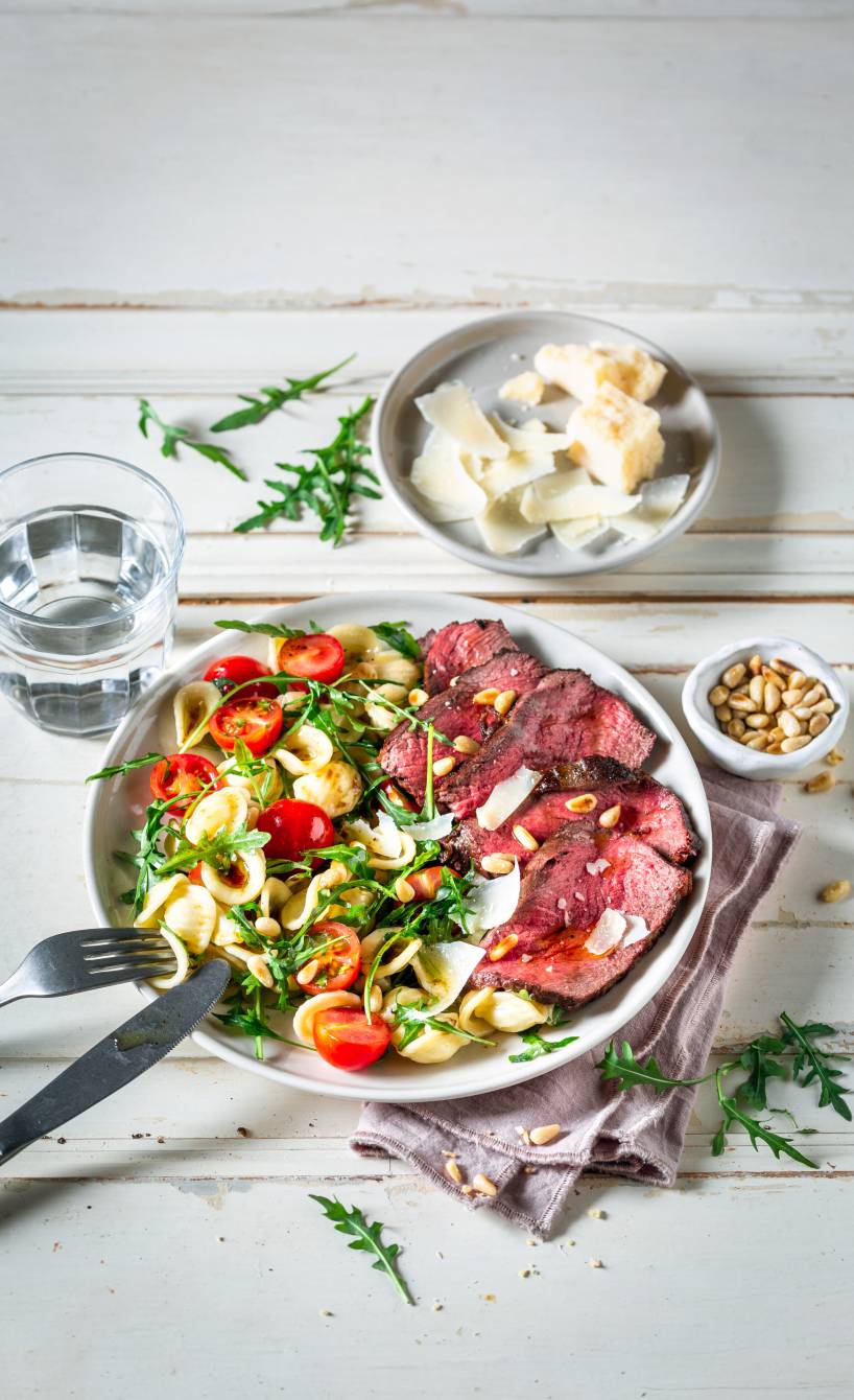 Teller mit Pastasalat auf der einen Seite und Rindfleischscheiben auf der anderen, daneben Parmesan und Pinienkerne auf einem Tisch