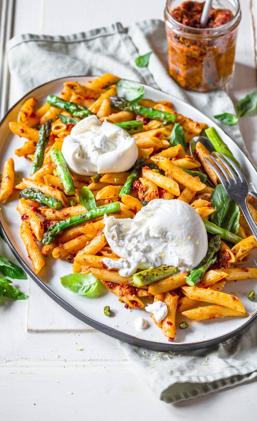 Nudelsalat aus Penne mit grünem Spargel auf einem ovalen Teller getoppt von Burratakugeln