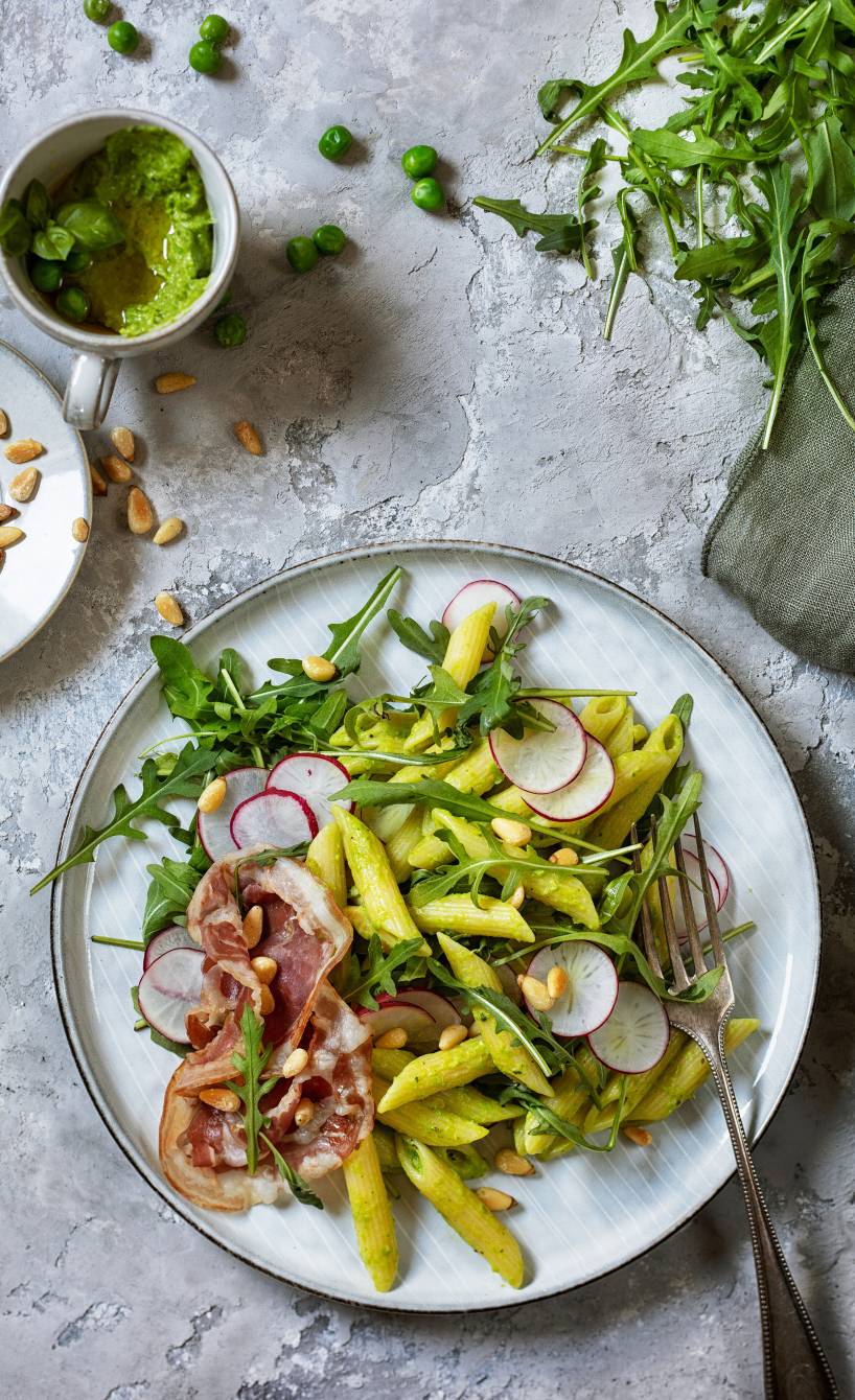 Penne-Salat auf einem Teller mit Pancettastreifen und Rucola, daneben grünes Pesto in einem Glas