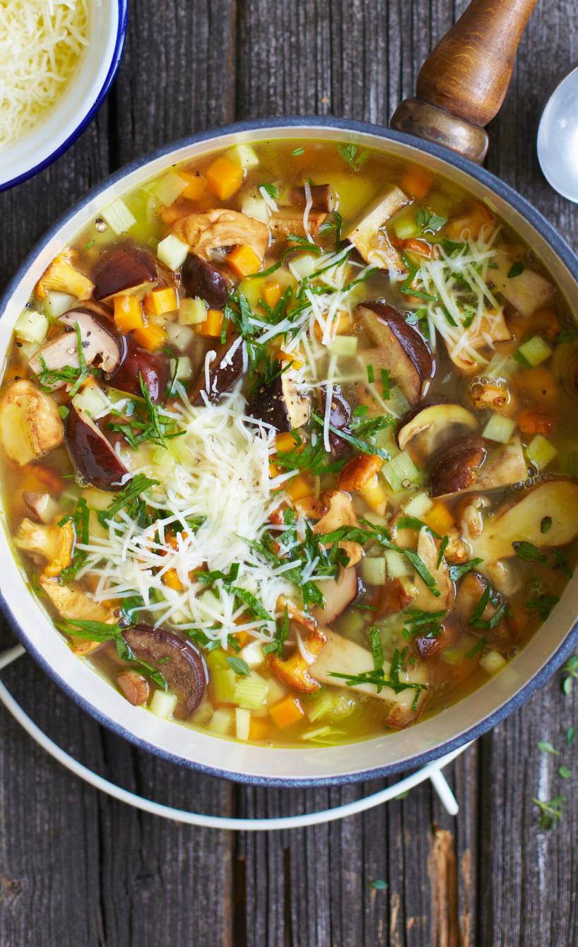 Klare Pilzsuppe mit Gemüse in einem Stieltopf auf einem Tisch, daneben gehobelter Käse und Baguette