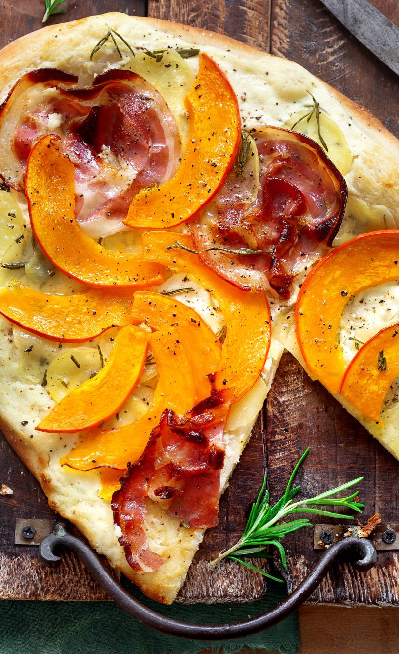 Pizza bianca mit Kürbisspalten, Kartoffeln und Speck belegt auf einem Holzbrett