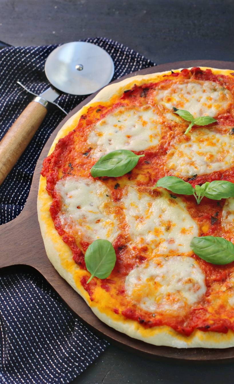 Runde, ganze Pizza Margherita auf einem Pizzabrett, daneben frische Basilikum in einem Topf