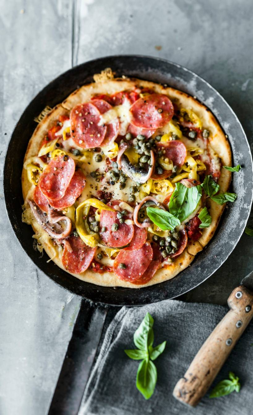 Pizza in einer Pfanne belegt mit Salami, Peperoni, Kapern und Sardellen