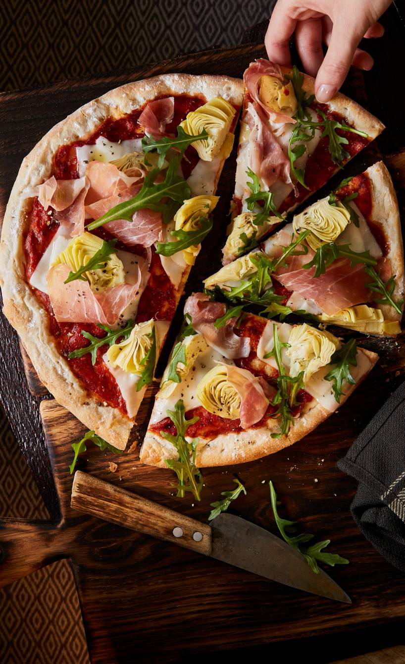 Runde Pfannenpizza mit Blag aus Schinken, Artischocken und Rucola angeschnitten auf einem Holzbrett