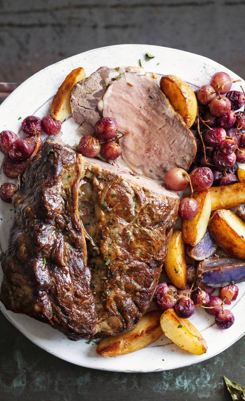 Gebackenes Roastbeef am Stück angeschnitten auf einem Teller umgeben von gebackenen Kartoffeln und Trauben