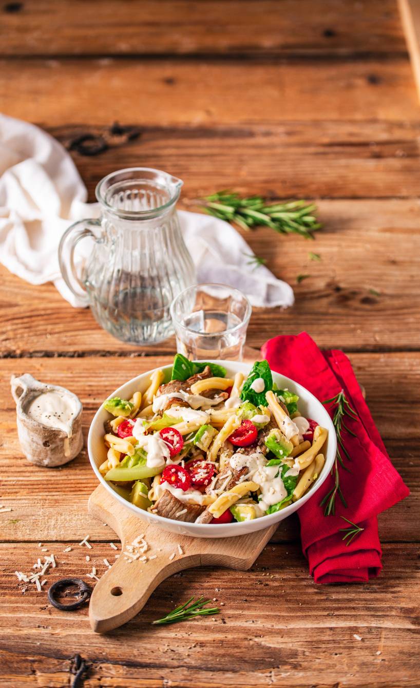 Pastasalat mit Romana auf einem Teller, getoppt von Kalbsschnitzelstreifen, Avocadostücken und einem cremigen Dressing