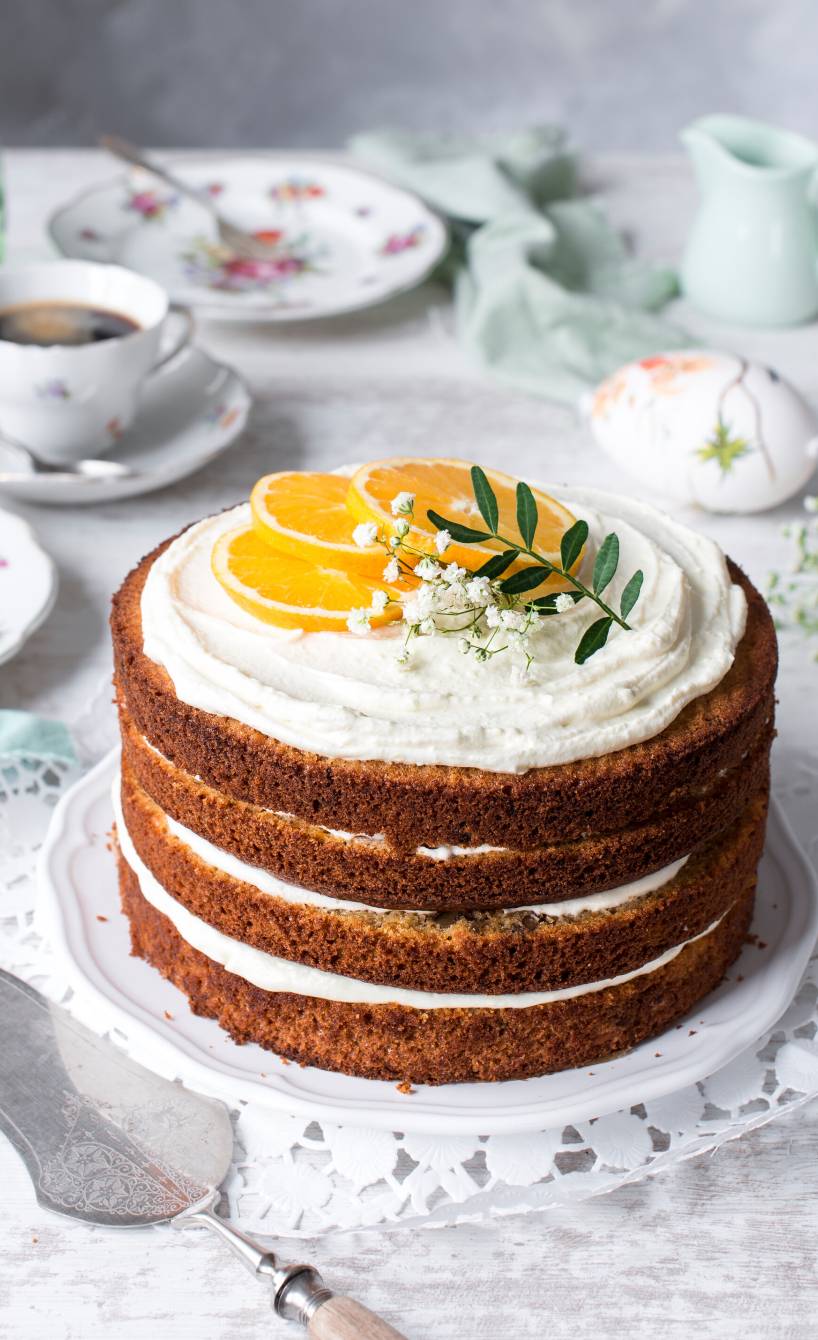 Rüblitorte mit Orangen-Frischkäse-Füllung getoppt von Orangenscheiben auf einer Tortenplatte auf einem österlich dekorierten Tisch