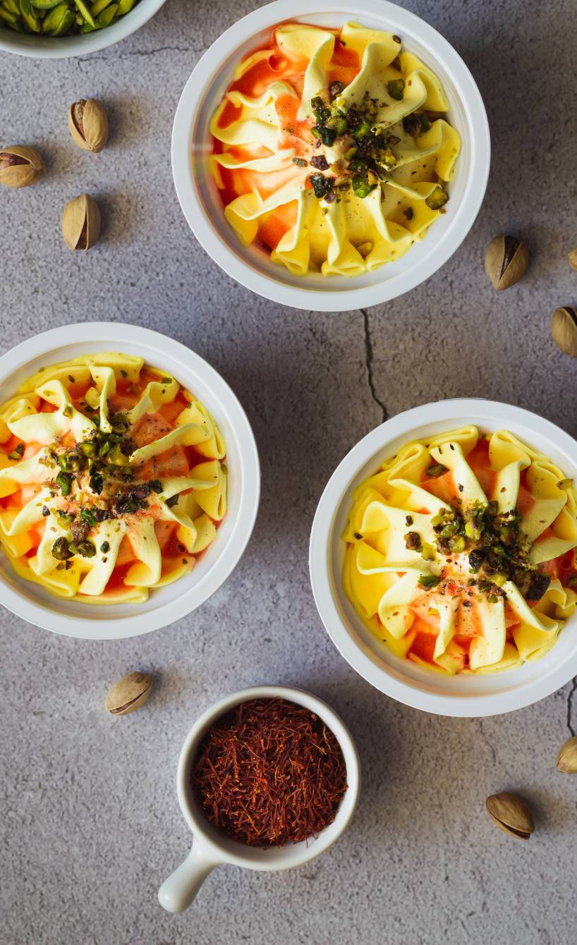 Selbstgemachtes Safran-Eis in drei Dessertschalen auf einem Tisch, daneben Safranfäden und Pistazienkerne