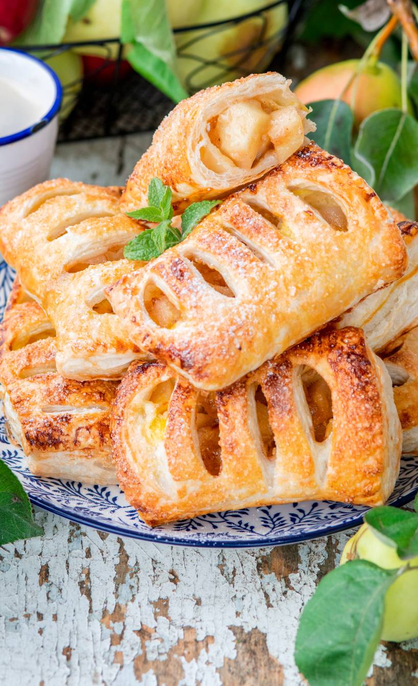 Bratapfeltaschen aus Blätterteig gestapelt auf einem Teller, daneben frische Äpfel auf einem Tisch