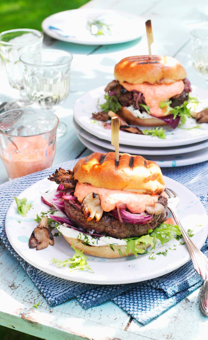 Gegrillter Burger mit Ziegenkäse und Pilzen gefüllt, zusammengehalten von einem Holzstab auf einem Teller, daneben ein Glas Mayonnaise