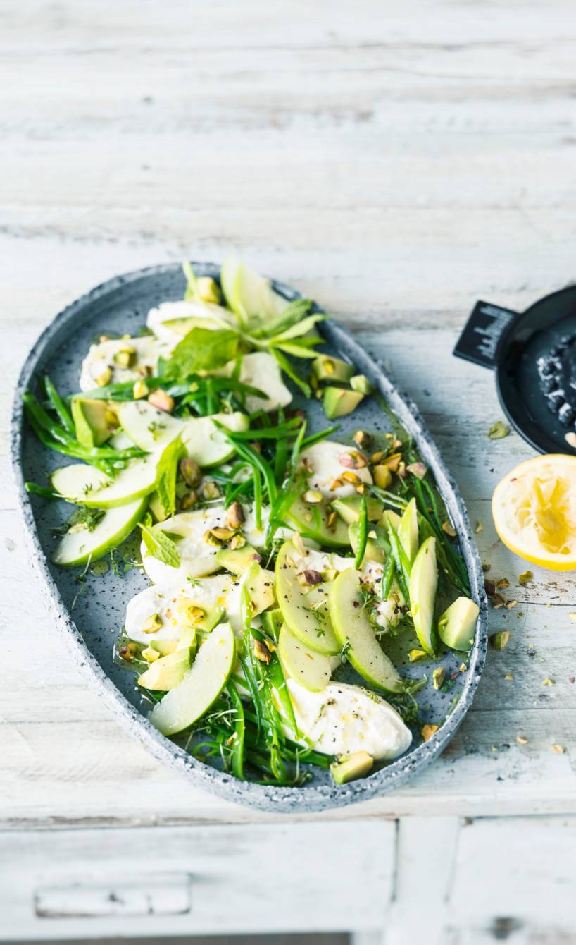 Burrata-Salat mit grünen Apfelspalten auf einem schwarzen Teller getoppt von Pistazien, daneben eine halbe Zitrone auf einem Tisch