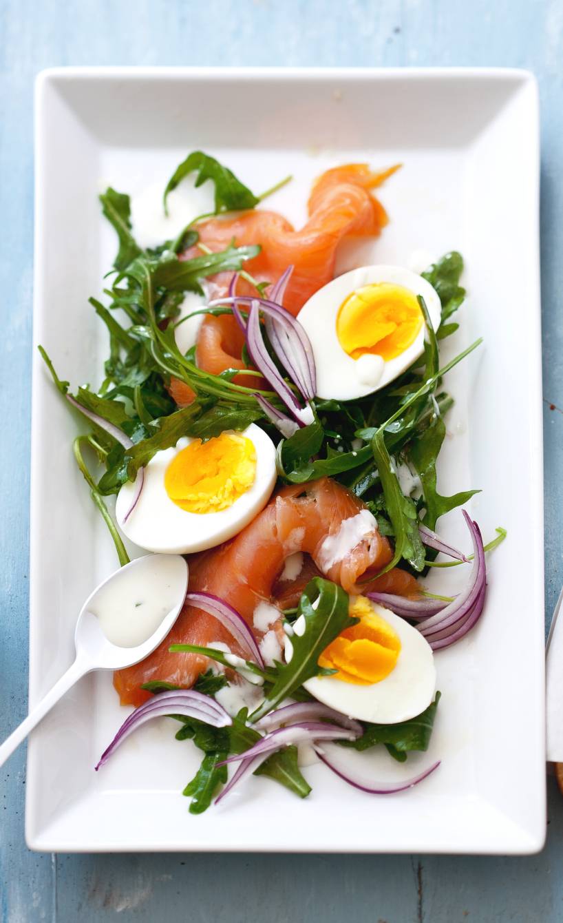 Rucola-Salat getoppt von RĂ€ucherlachsscheiben, halben gekochten Eiern und Joghurt-Dressing angerichtet auf einer Salatplatte