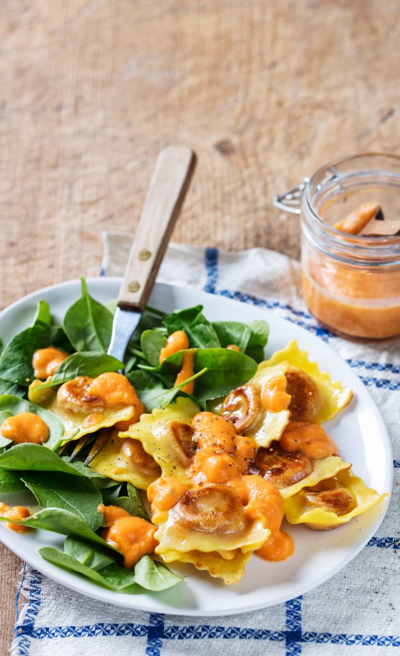 Gebratene Ravioli neben Babyspinat auf einem Teller, daneben Karottensoße in einem Weckglas