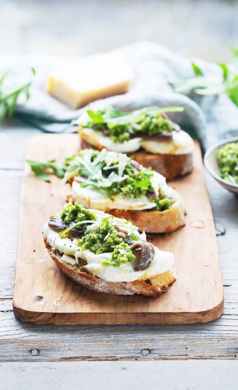 Crostini auf einem Holzbrett getoppt von Sardellenfilets, Mozzarella und grünem Pesto