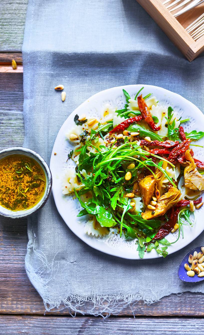 Farfalle-Salat angerichtet auf einem Teller mit Artischocken, Rucola, getrockneten Tomaten und Pinienkernen