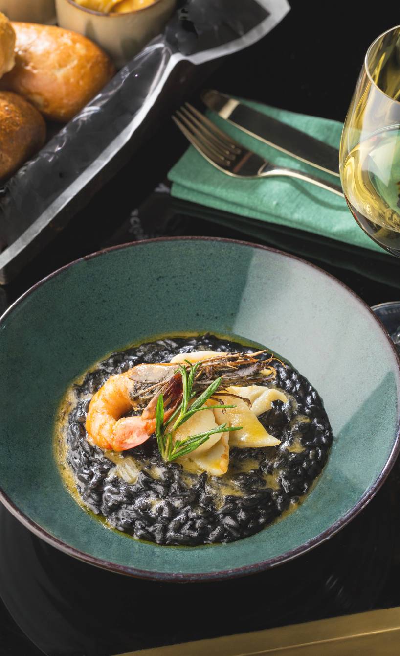 Schwarzes Meeresfrüchterisotto auf einem tiefen Teller garniert mit Kräutern und Garnelen, daneben ein Glas Wein auf einem Tisch