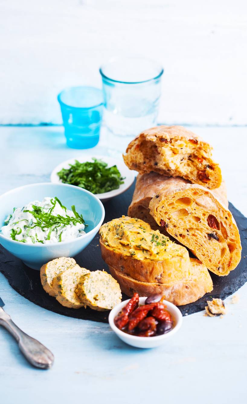 Selbstgebackenes Ciabatta mit getrockneten Tomaten und Oliven in Scheiben auf einem Teller, daneben Bärlauchbutter und Ricottacreme