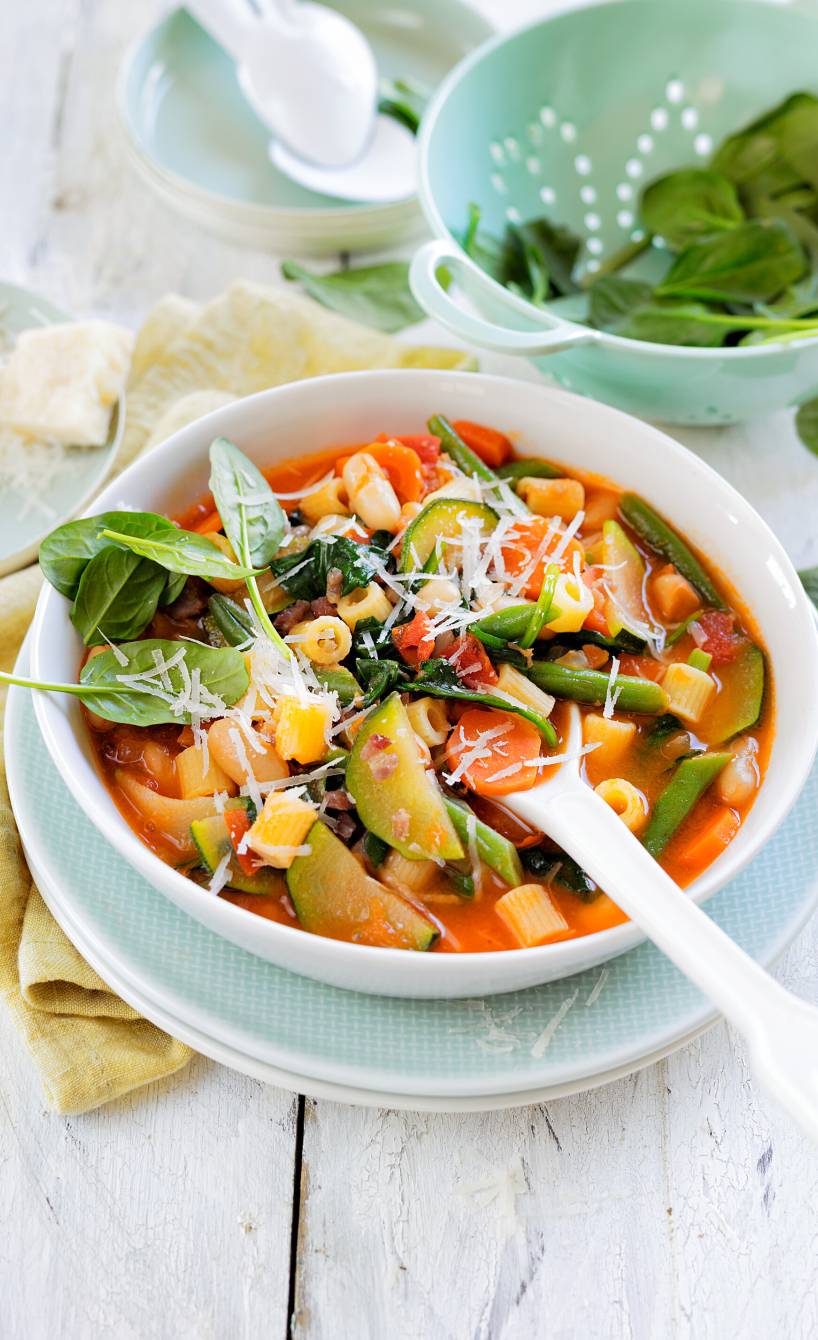 Sommer-Minestrone in einer Suppenschüssel, daneben Babyspinatblätter und Parmesan in Schalen