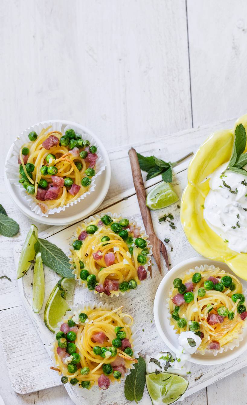 Mehrere Spaghetti-Muffins mit Schinken in papierförmchen auf einem Tisch, daneben Minz-Limetten-Dip