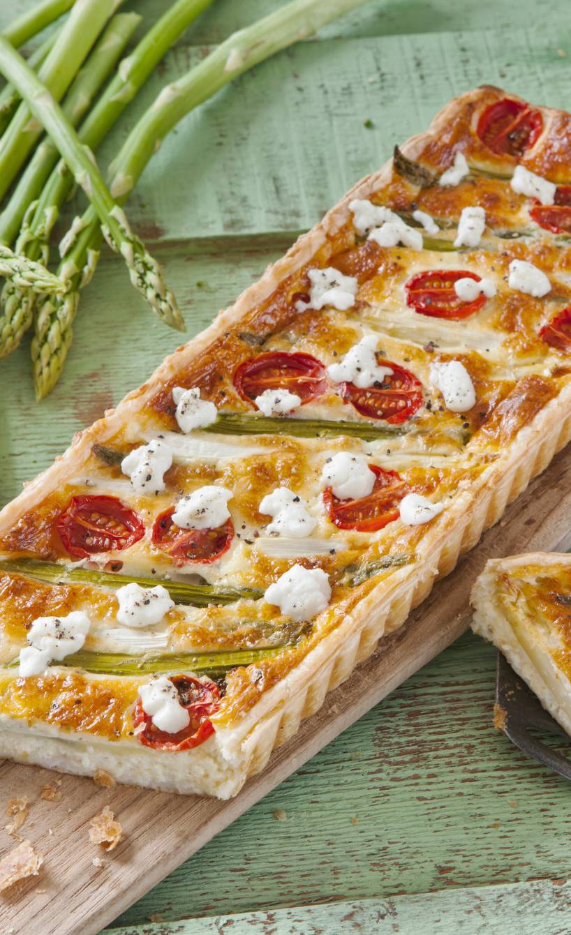 Reechteckige Spargeltarte mit Ziegenfrischkäse und Kirschtomaten angeschnitten auf einem Holzbrett