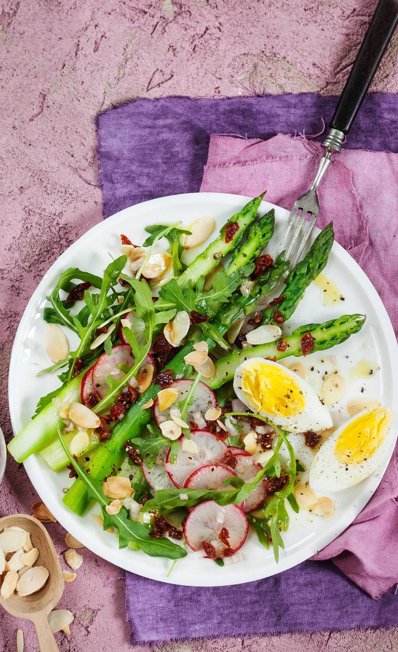 Salat aus grünem Spargel, Radieschen und Rucola auf einem Teller getoppt mit Ei, daneben Dressing