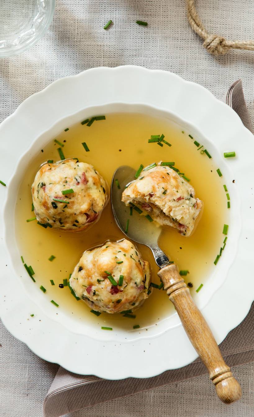 Südtiroler Speckknödel in einem Suppenteller mit Brühe, daneben frischer Schnittlauch auf einem Holzbrett