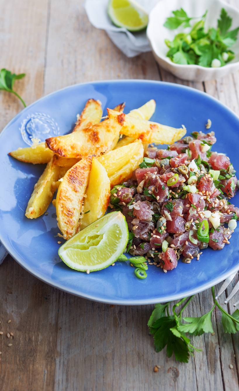 Thunfisch-Tartar angerichtet auf einem Teller, daneben Parmesankartoffelspalten, Limette und Petersilie