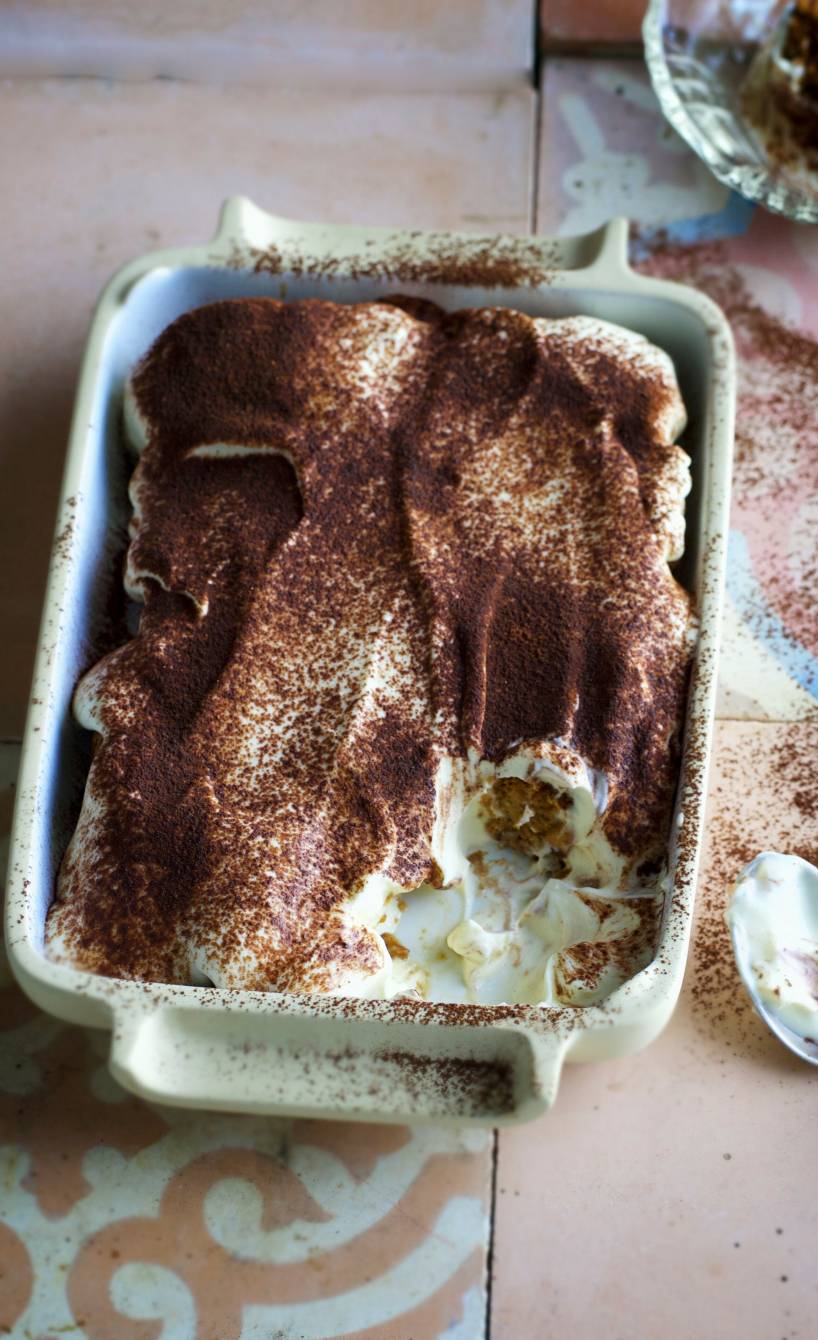 Tiramisu in einer Auflaufform mit einem kleinen fehlenden teil, daneben ein benutzter Löffel auf einem Tisch