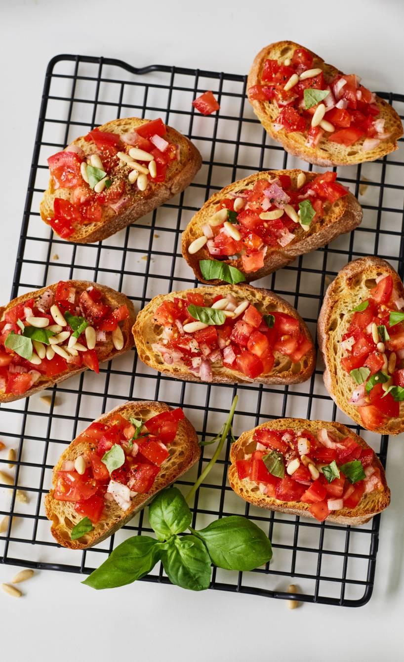 Geröstete Ciabattascheiben belegt mit Tomaten, Zwiebeln, Pinienkernen und Basilikum auf einem Backgitter